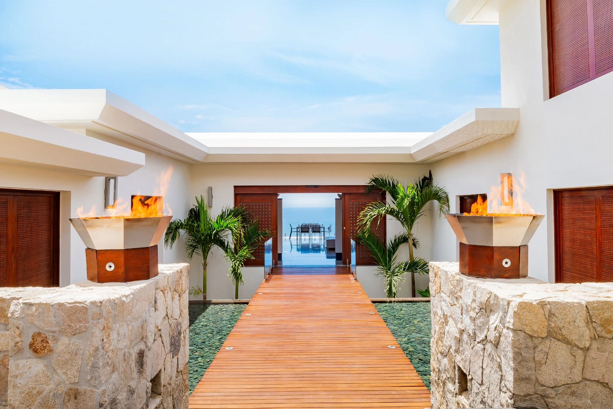 Villa entrance with a timber walkway, palms and fire bowls framing the ocean view